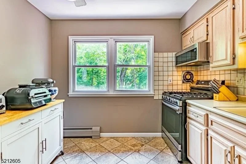 Kitchen with beige cabinets, tan tiled floor, beige walls, and a large window overlooking green trees. Appliances include a microwave, stove, and toaster on counters.