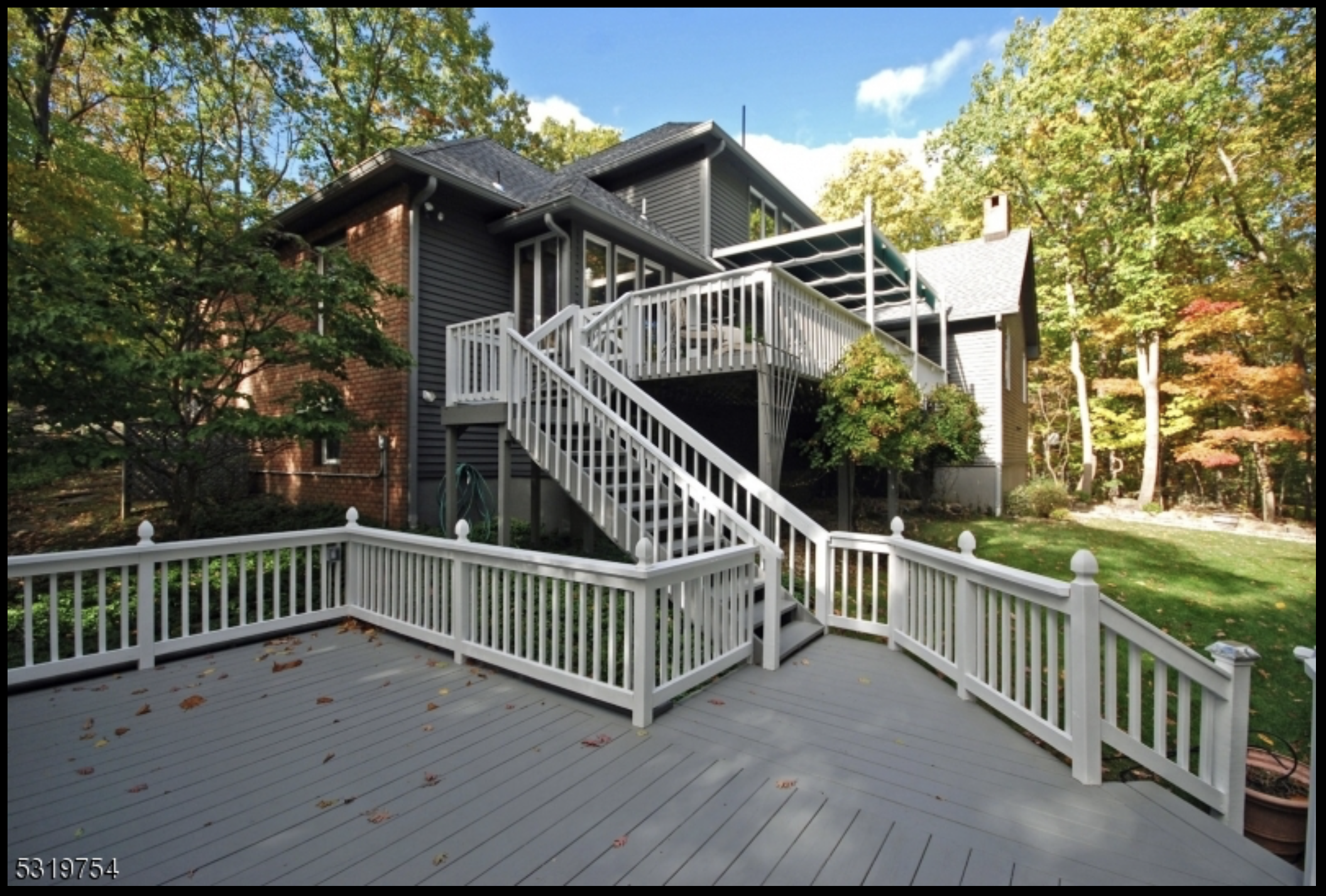 A large multi-level wooden deck attached to a dark gray house, surrounded by trees with autumn foliage. The deck has white railings and a staircase leading down to a grassy area. There's a garden hose hanging on the side of the house.