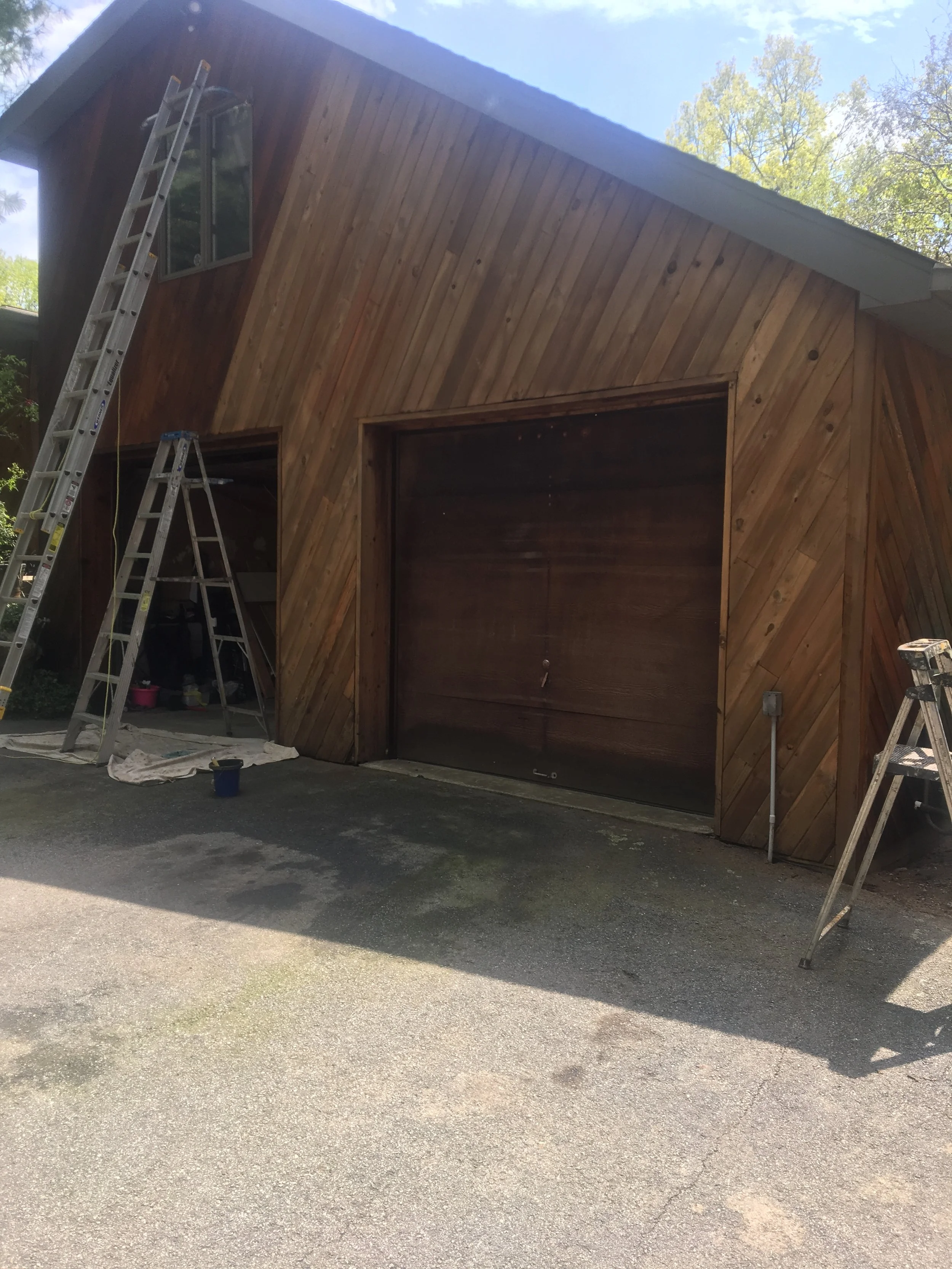 Wooden garage door with diagonally arranged wood paneling, part of a building under construction with ladders leaning against it, and construction tools and materials nearby.