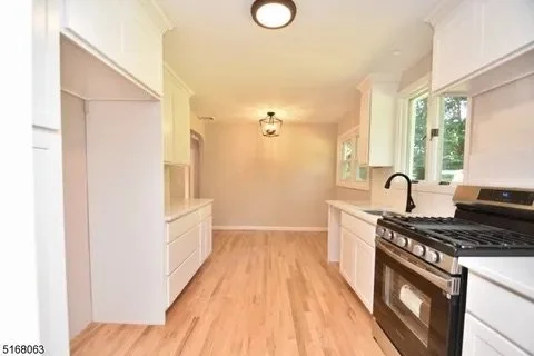 Empty kitchen with white cabinets, a black stove, and wooden flooring, illuminated by ceiling and window light.