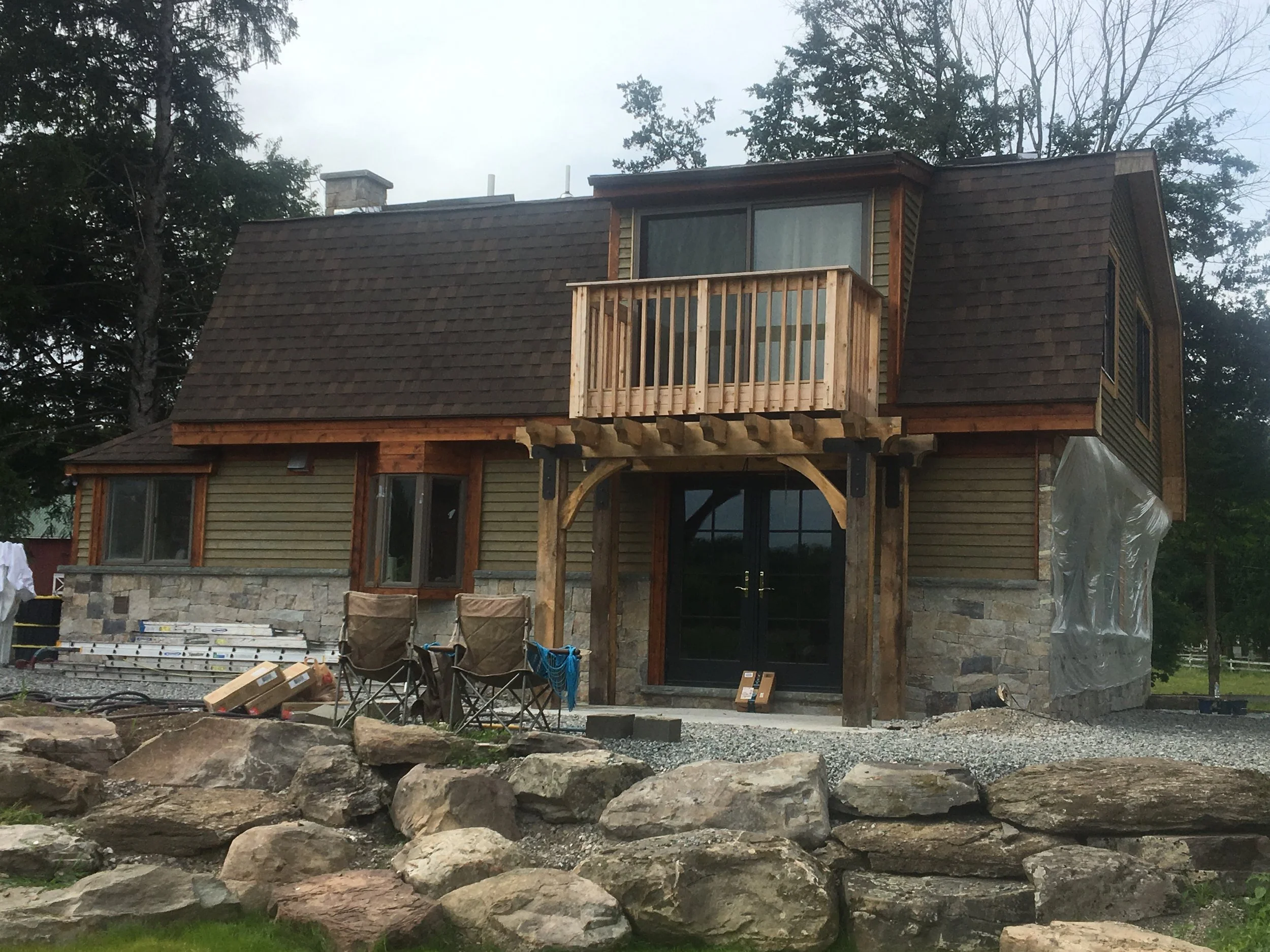 A house under construction with a stone foundation, wooden siding, and a new balcony on the second floor. There are construction chairs and materials in front, and large rocks along the front yard.