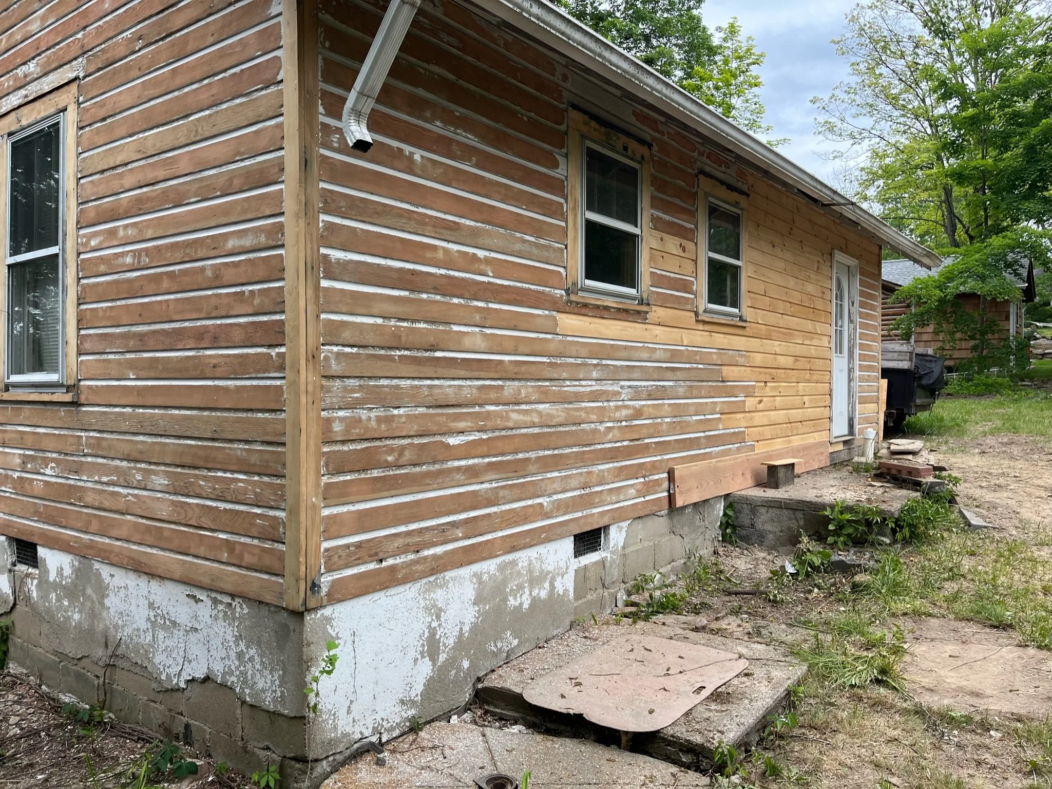 A house exterior under renovation showing partially replaced wooden siding, some sections with weathered siding and others with new wood planks, with a concrete block foundation and a small concrete step in front of a door.