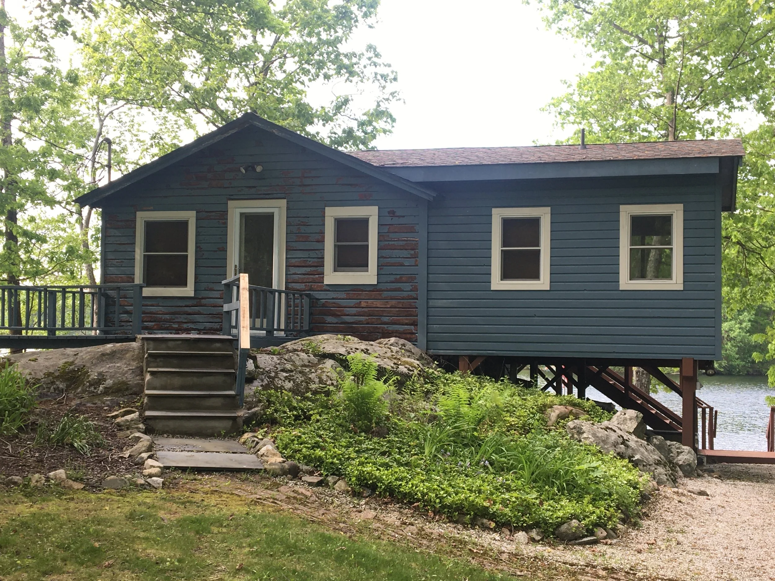 A small, weathered blue wooden house on a rocky hill by a lake, surrounded by green trees, with shrubbery in the front and stairs leading to a small deck.