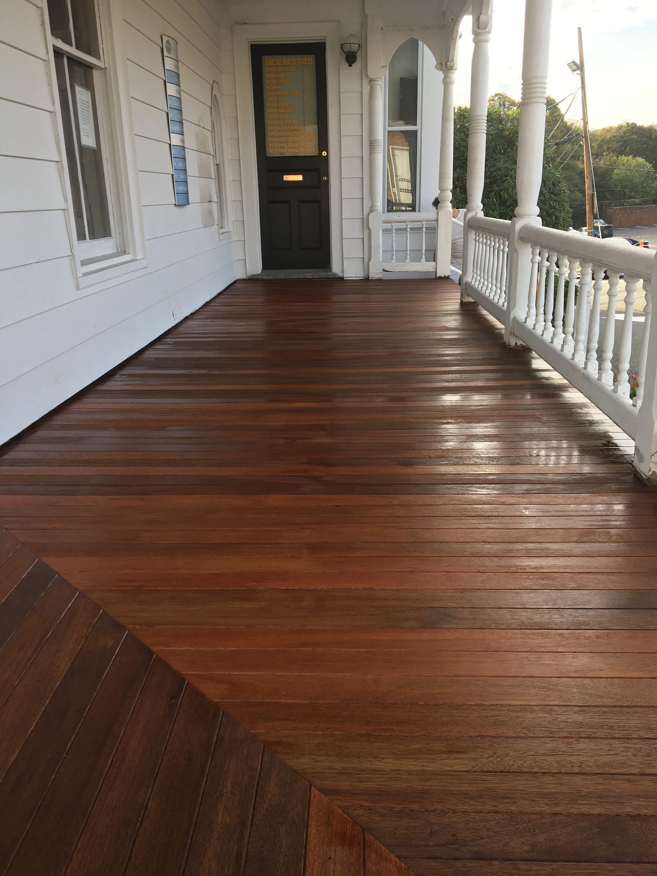 Wooden porch with white railing in front of a black door on a white house, with a tree and power pole in the background.