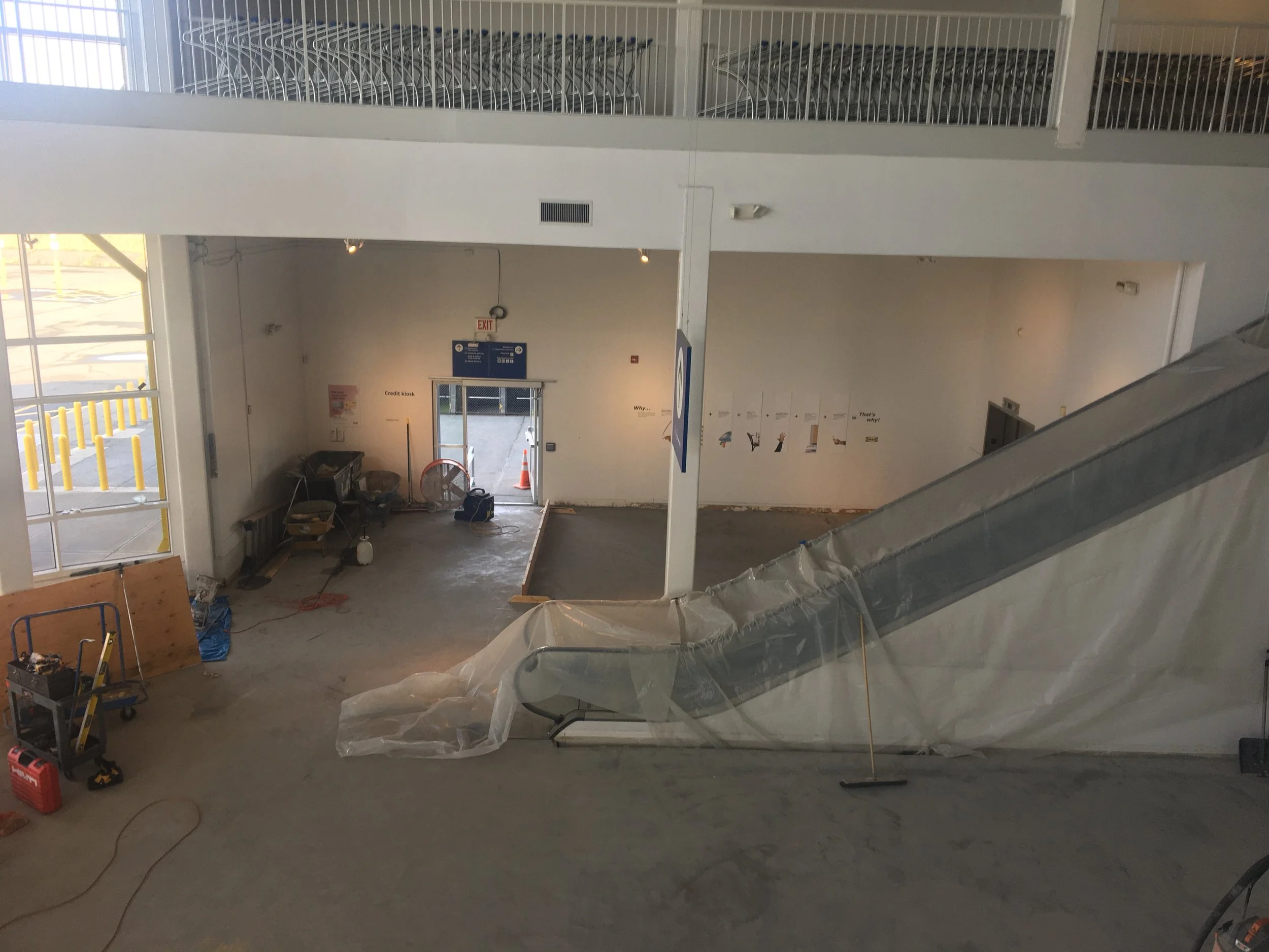 Interior view of a building under construction with an escalator partially covered in plastic, construction tools and materials scattered on the floor, and construction signs on the wall.