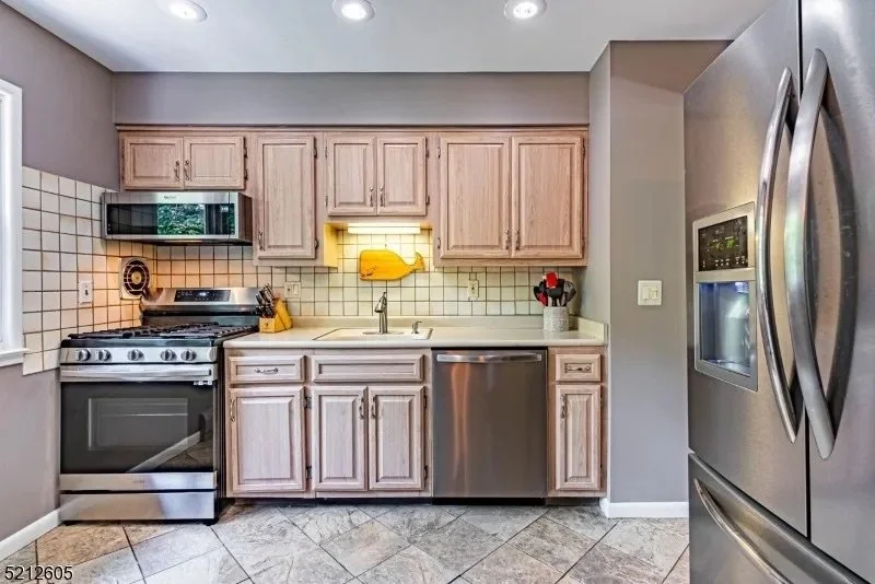 Kitchen with light wood cabinets, stainless steel dishwasher, oven, and refrigerator, white tiled backsplash, gray wall, and tiled floor.