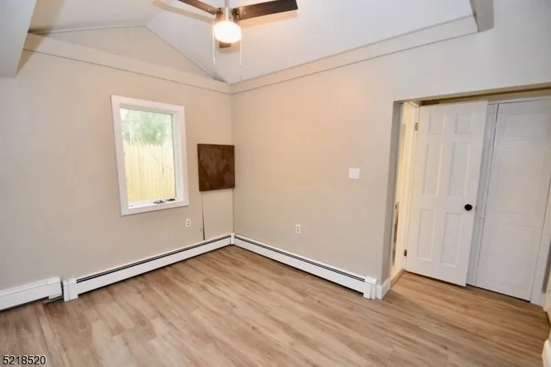 Empty room with beige walls, a window, a ceiling fan with a light, a door leading to another room, and light wood flooring.