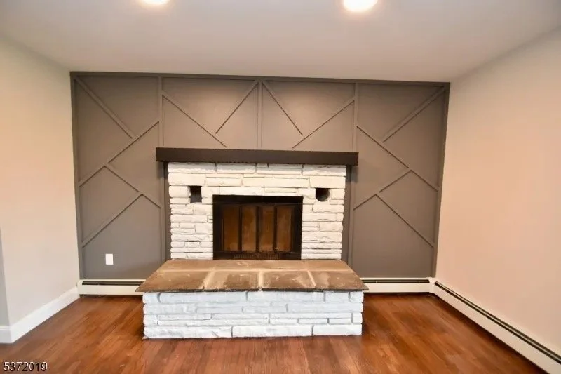 Living room with a white brick fireplace and a geometric gray wall behind it, hardwood floor, and baseboard heating.