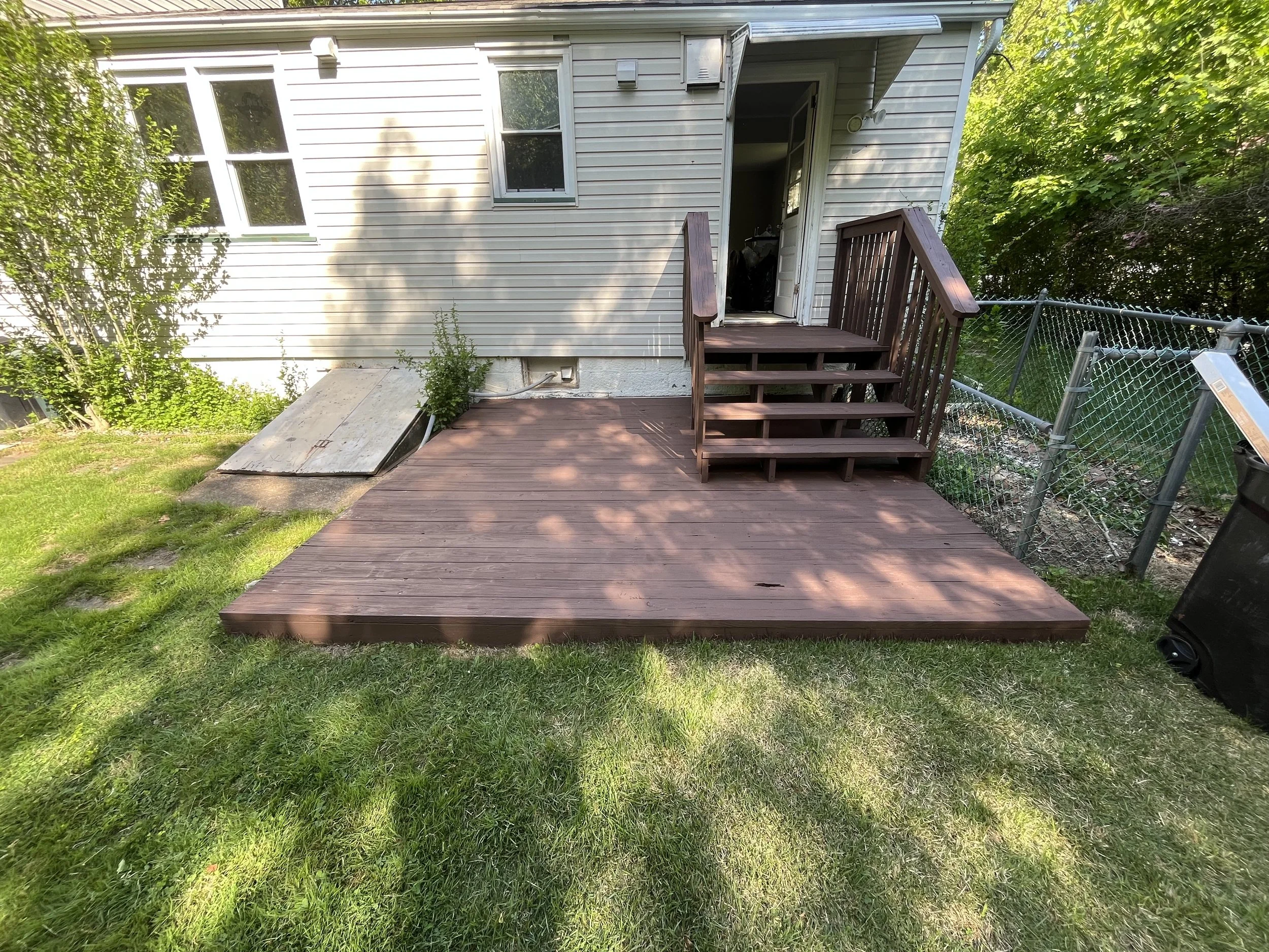 Backyard with a newly built wooden deck attached to a house, with steps leading to a sliding door, surrounded by green grass, trees, and a chain-link fence.
