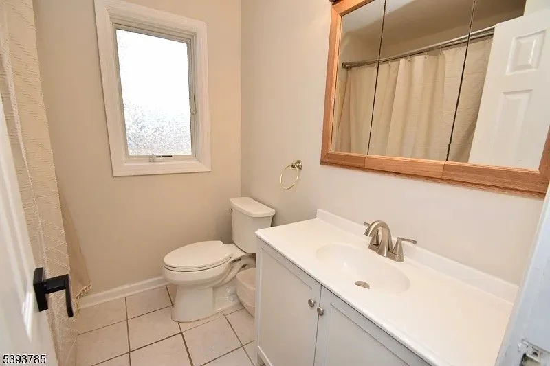 A small bathroom with a toilet, a white vanity with a sink, a mirror, a window, and a shower with a curtain.