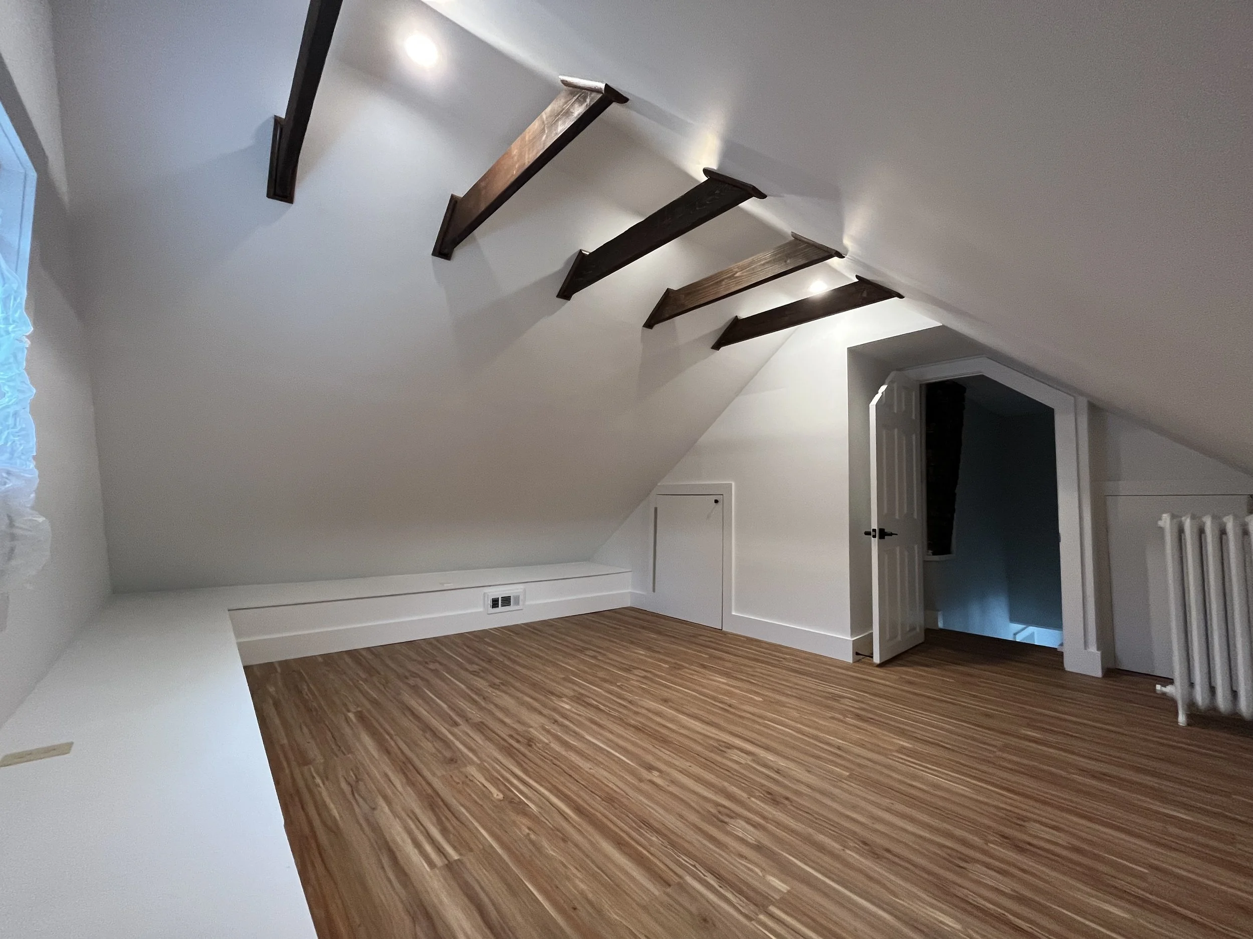 Empty attic room with sloped ceiling, wood beams, hardwood floor, white walls, small door on the wall, open doorway leading downstairs, window on the side, and radiator.