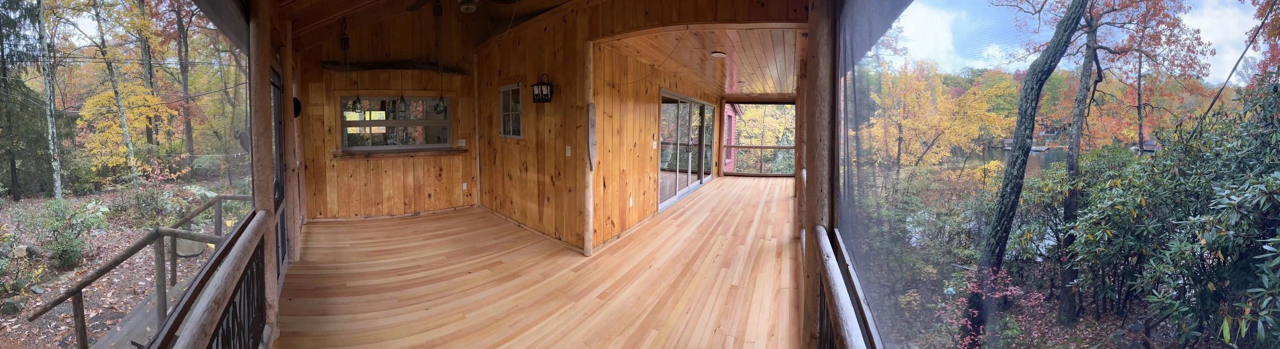 Interior view of a wooden house balcony or porch with large screened windows showing colorful fall trees outside.