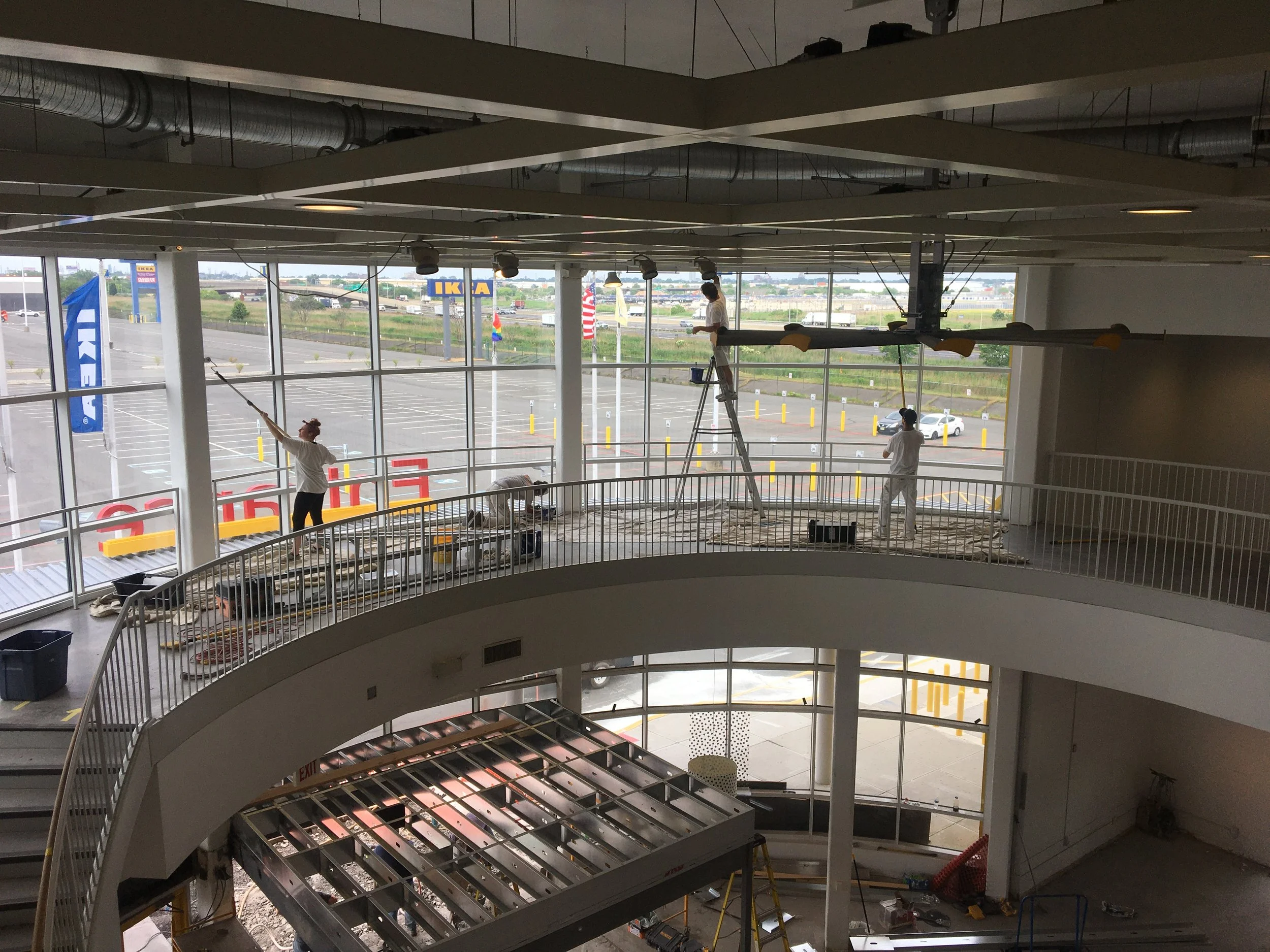 Workers cleaning and renovating the interior of a large commercial space, with large windows, a staircase, and suspended ceiling grid.