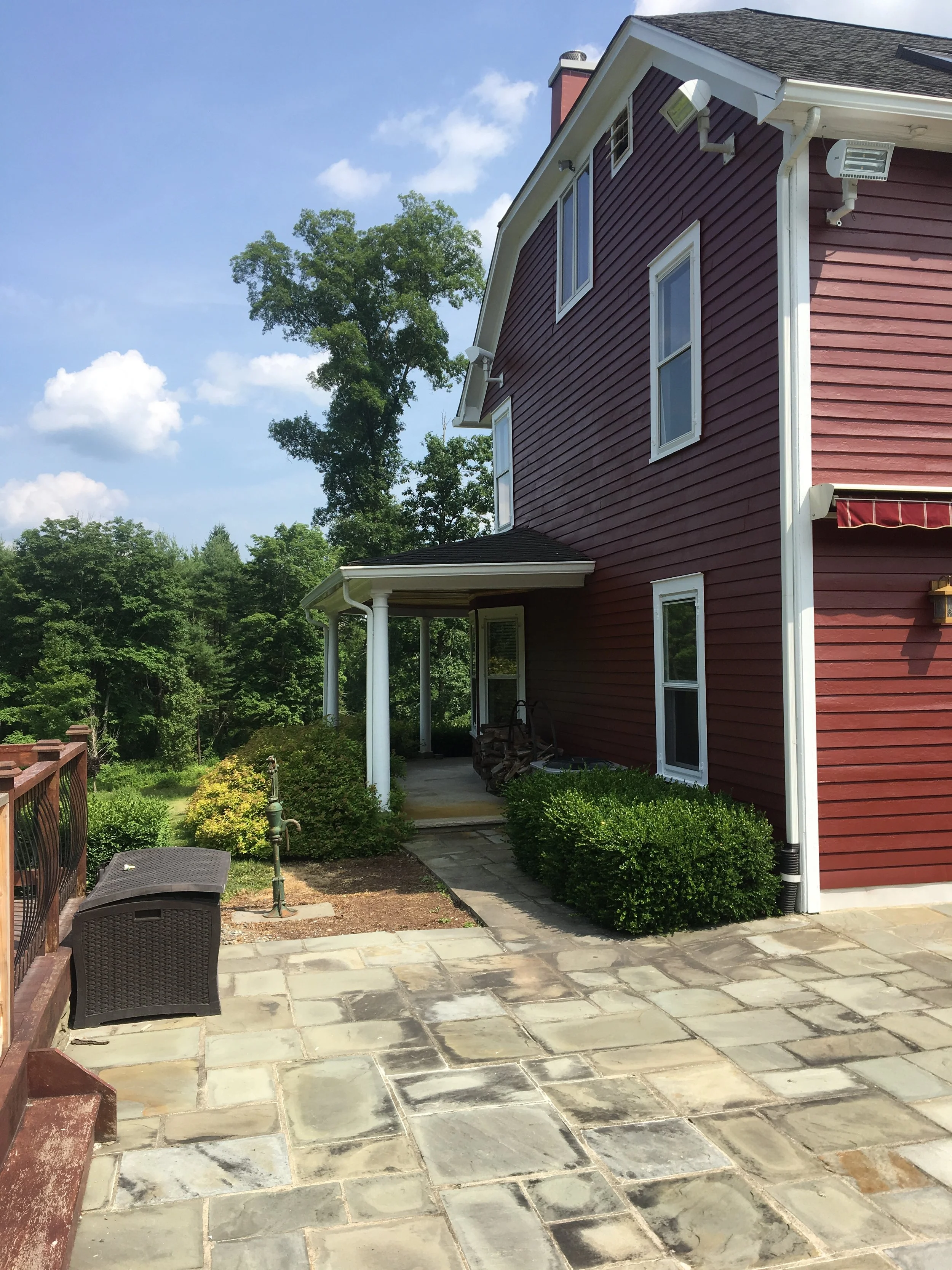 Backyard patio with stone pavers, a small garden bed, bushes, a wicker storage bench, a sprinkler, and the side of a red house with white trim, windows, and a covered porch. In the background, a large green tree and a blue sky with some clouds.