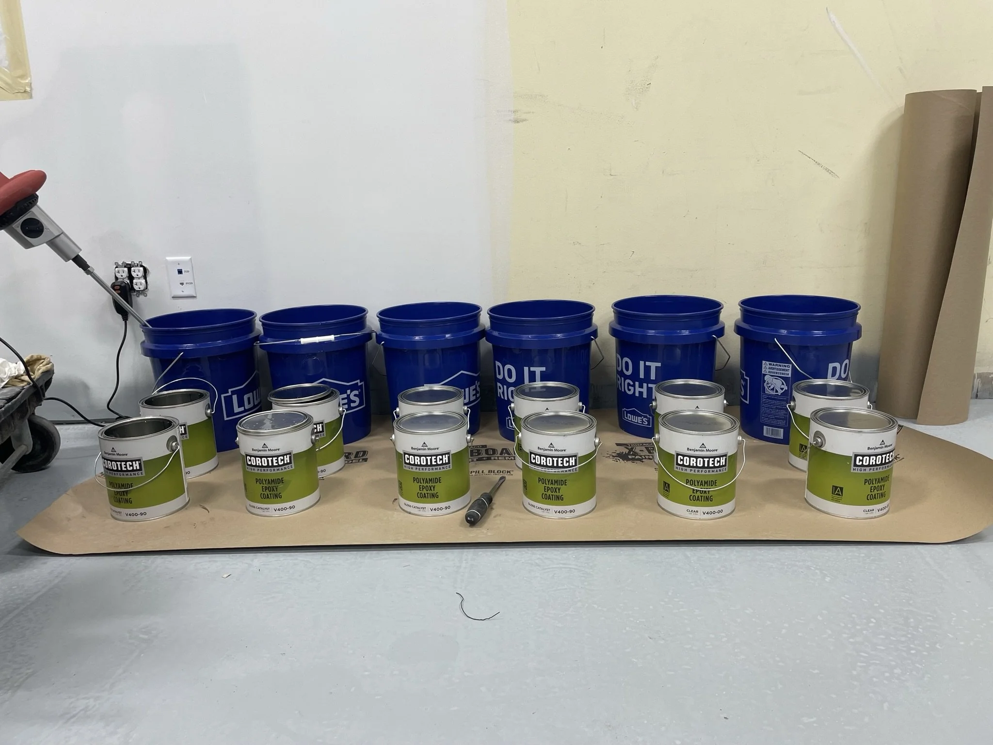 Several blue Lowe's buckets and multiple white cans of Corotech epoxy coating arranged on a piece of cardboard on a table in a workshop setting.