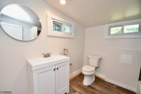 Bathroom with a white vanity, round mirror, and toilet, with wood flooring and small windows.