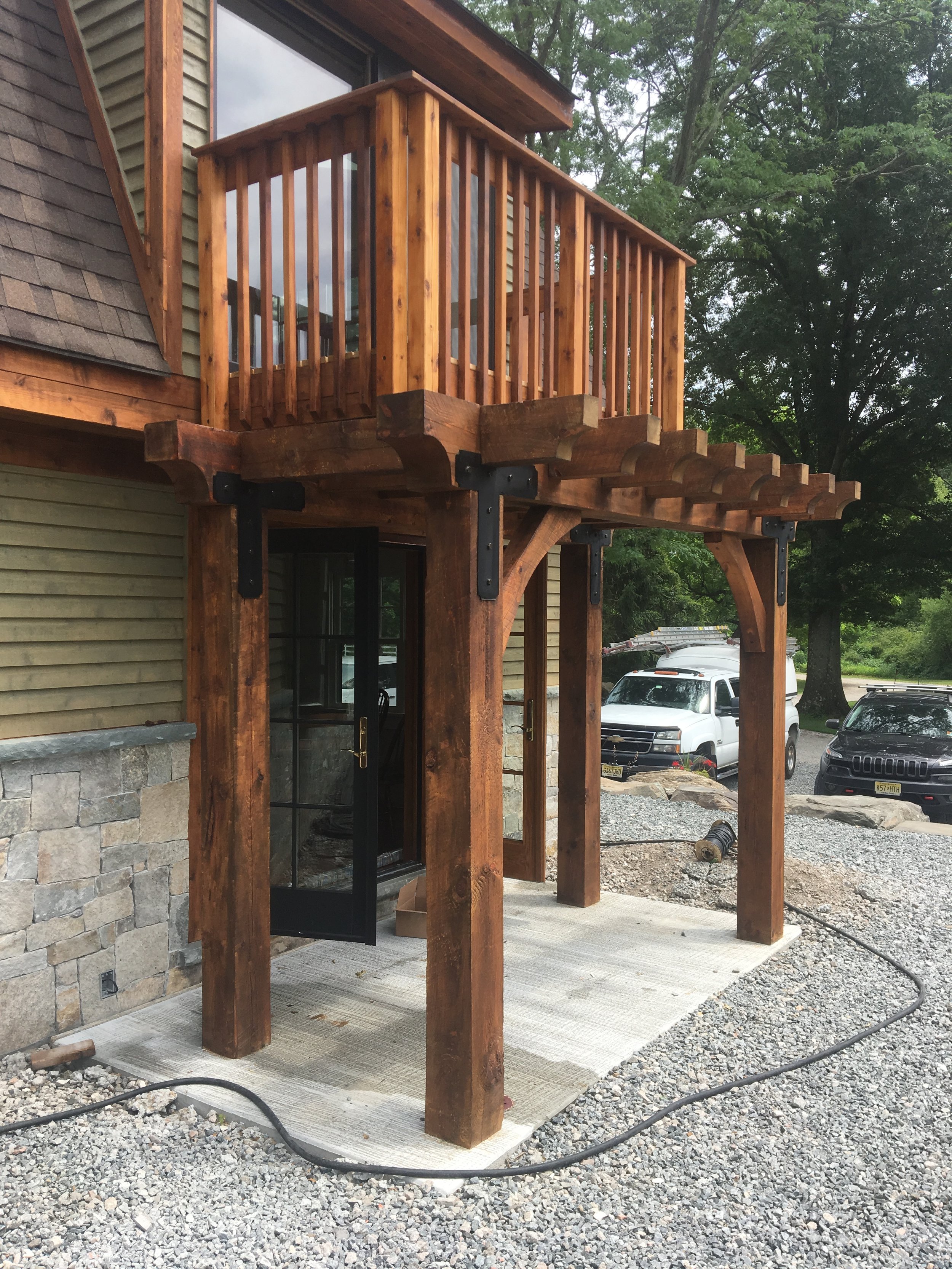Wooden balcony with railings above a covered patio area supported by large wooden posts on a gravel surface.