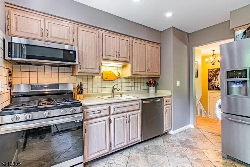 Kitchen with light wood cabinets, stainless steel appliances, and beige tile backsplash, leading to a hallway with a staircase and laundry area in the background.