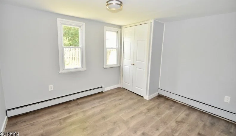 Empty room with light gray walls, two windows, white door, wood flooring, and baseboard heating.