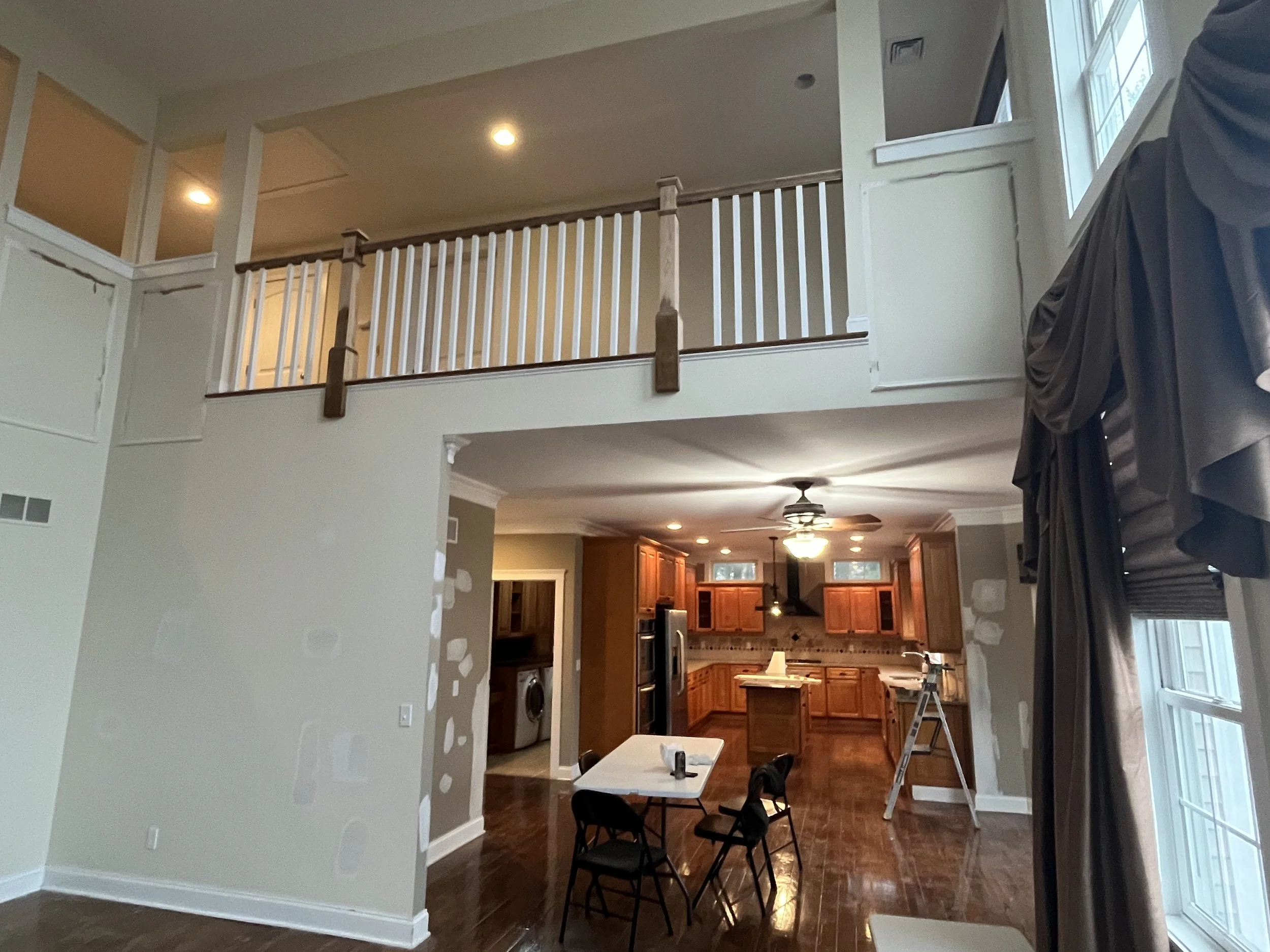 Interior view of a two-story home with a dining area, kitchen with wooden cabinets, and a laundry area with a washer and dryer in the background. There is a high ceiling with a ceiling fan, and a balcony with a white railing upstairs. A small table w