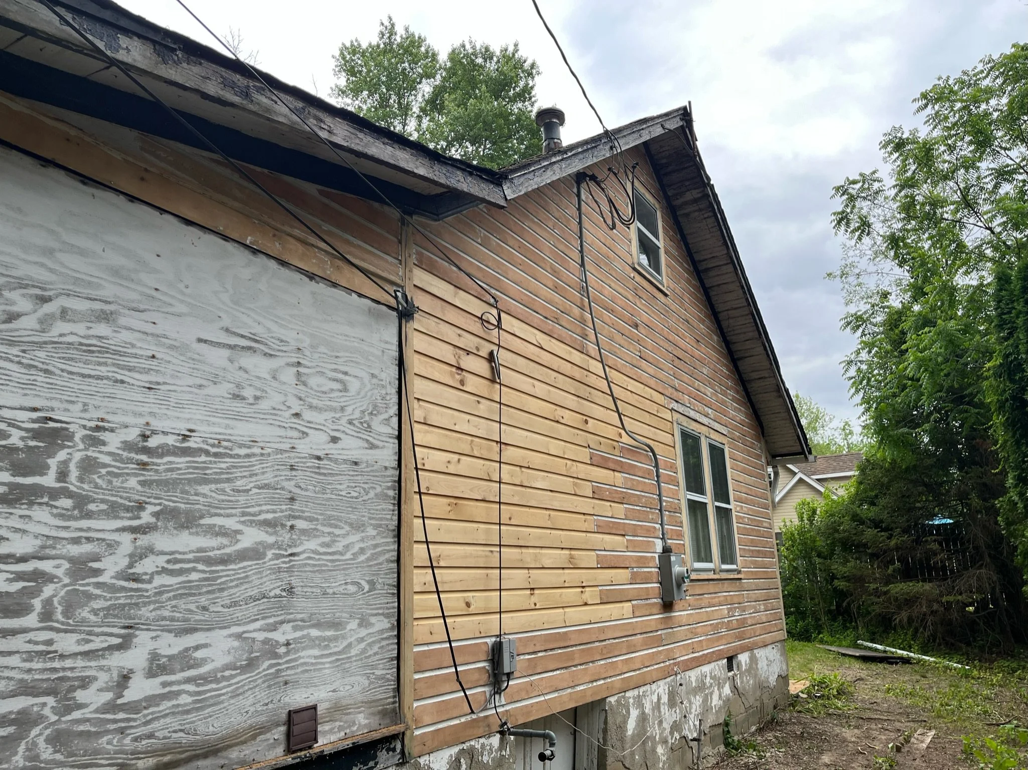 Side view of a house under renovation, with some exterior walls covered in weathered plywood and some in new wooden siding, trees, and cloudy sky in the background.