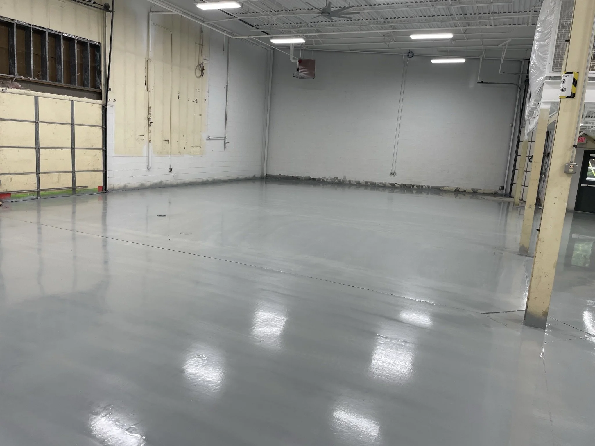 Empty, polished concrete floor in a large industrial or commercial space with unfinished sections on the wall and ceiling, and a partially open garage door on the left.