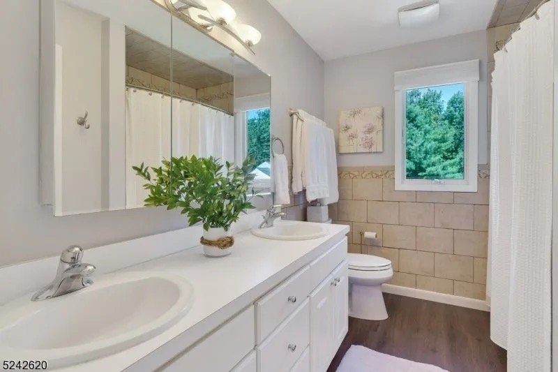 Bathroom with a double sink vanity, large mirror, potted plant, towel rack with white towels, window showing trees outside, and a shower curtain.