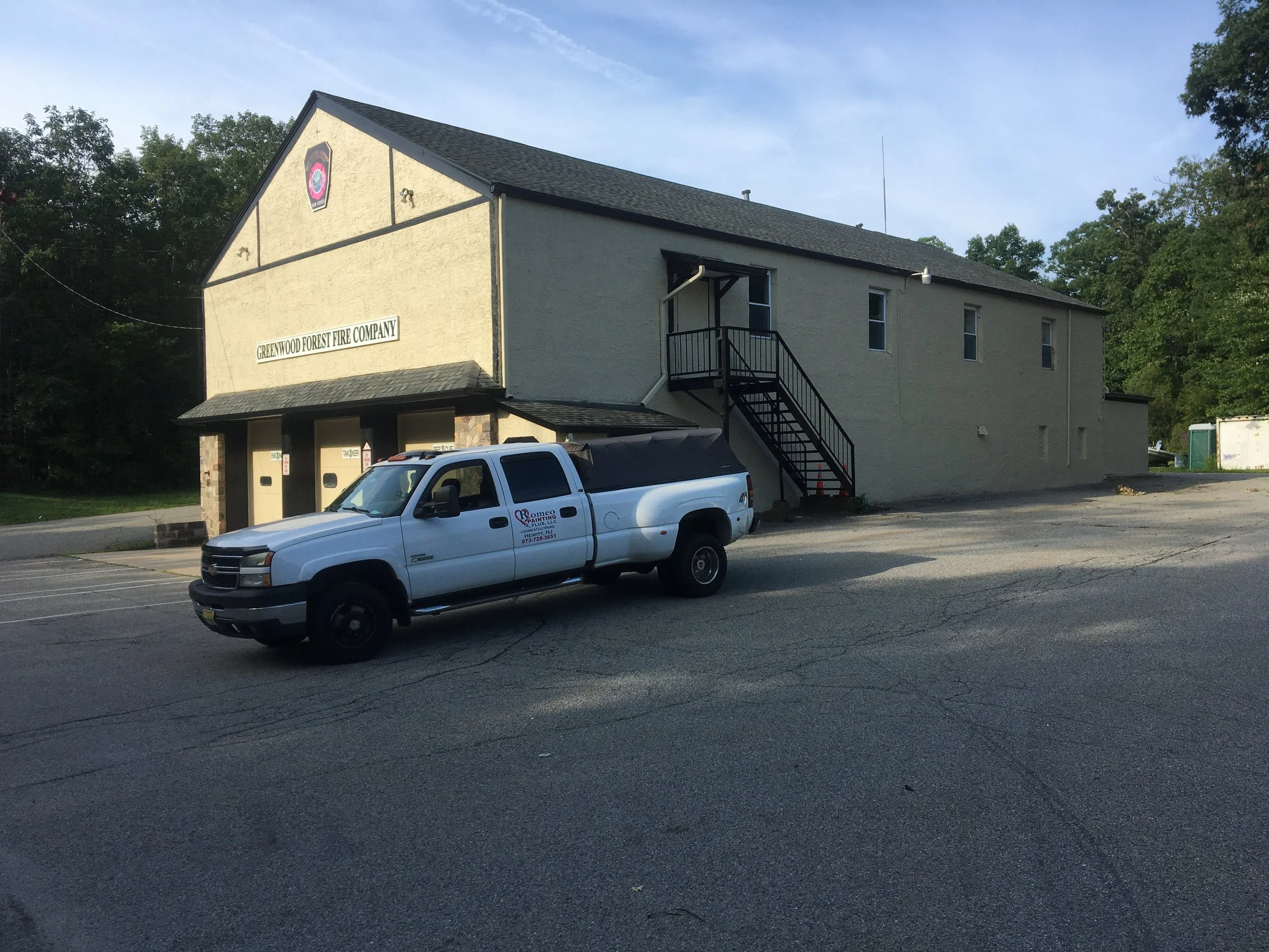 A two-story beige building with a sign that reads 'Greenwood Forest Fire Company' and an emergency vehicle parked in front.