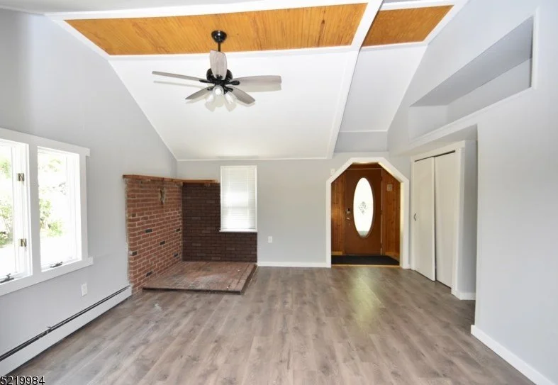 Empty living room with light gray walls, a brick fireplace, large window, dark wood flooring, and a ceiling fan, with an entryway with an oval glass door visible.