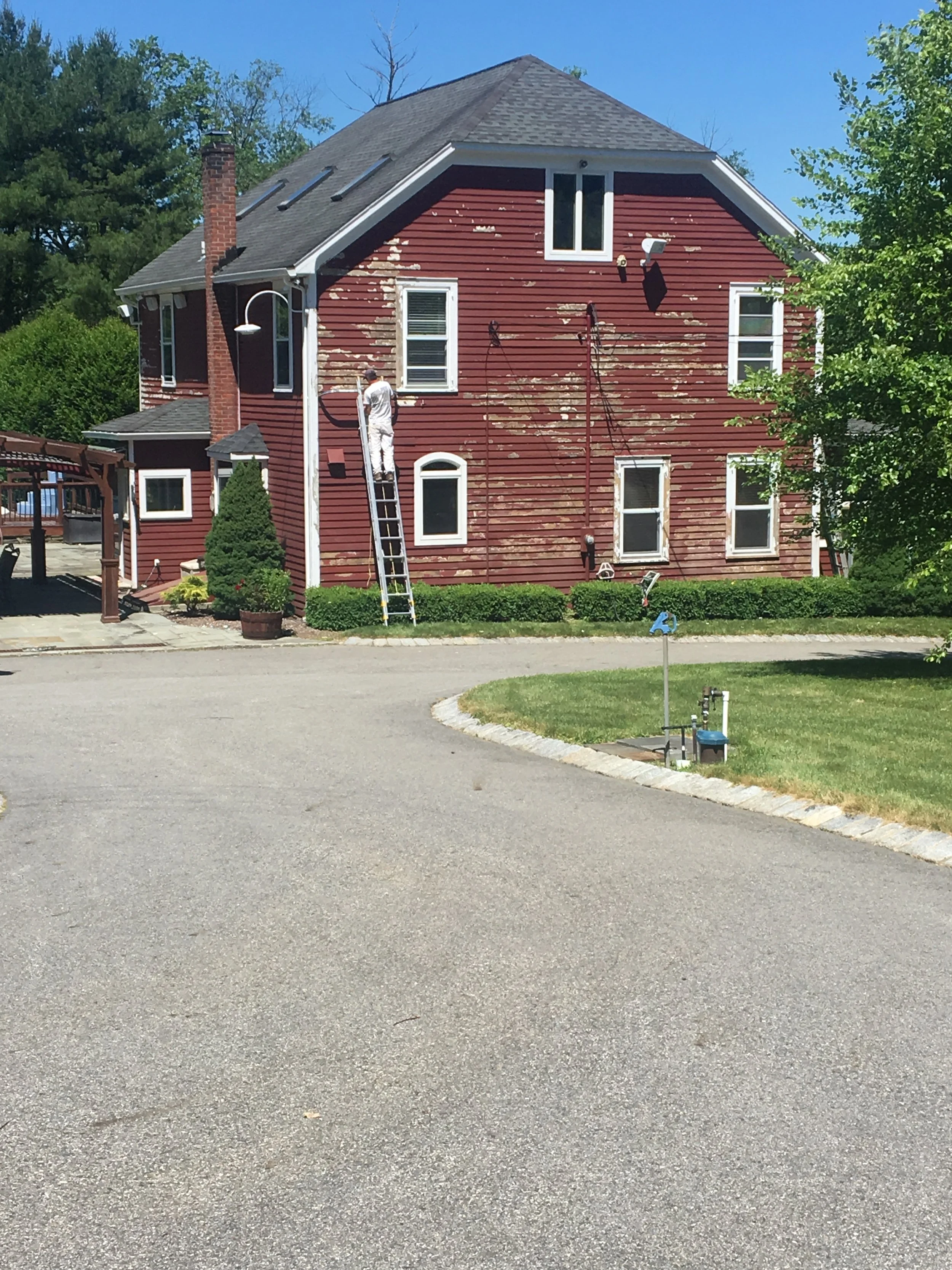 Worker painting the exterior of a red house with peeling paint, using a ladder and paint sprayer on a sunny day.