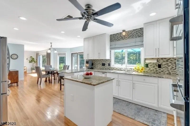 Bright open kitchen with white cabinets, a small island, and a mosaic tile backsplash. Connected to a dining area with a round table and chairs, large windows with blinds, and hardwood floors.