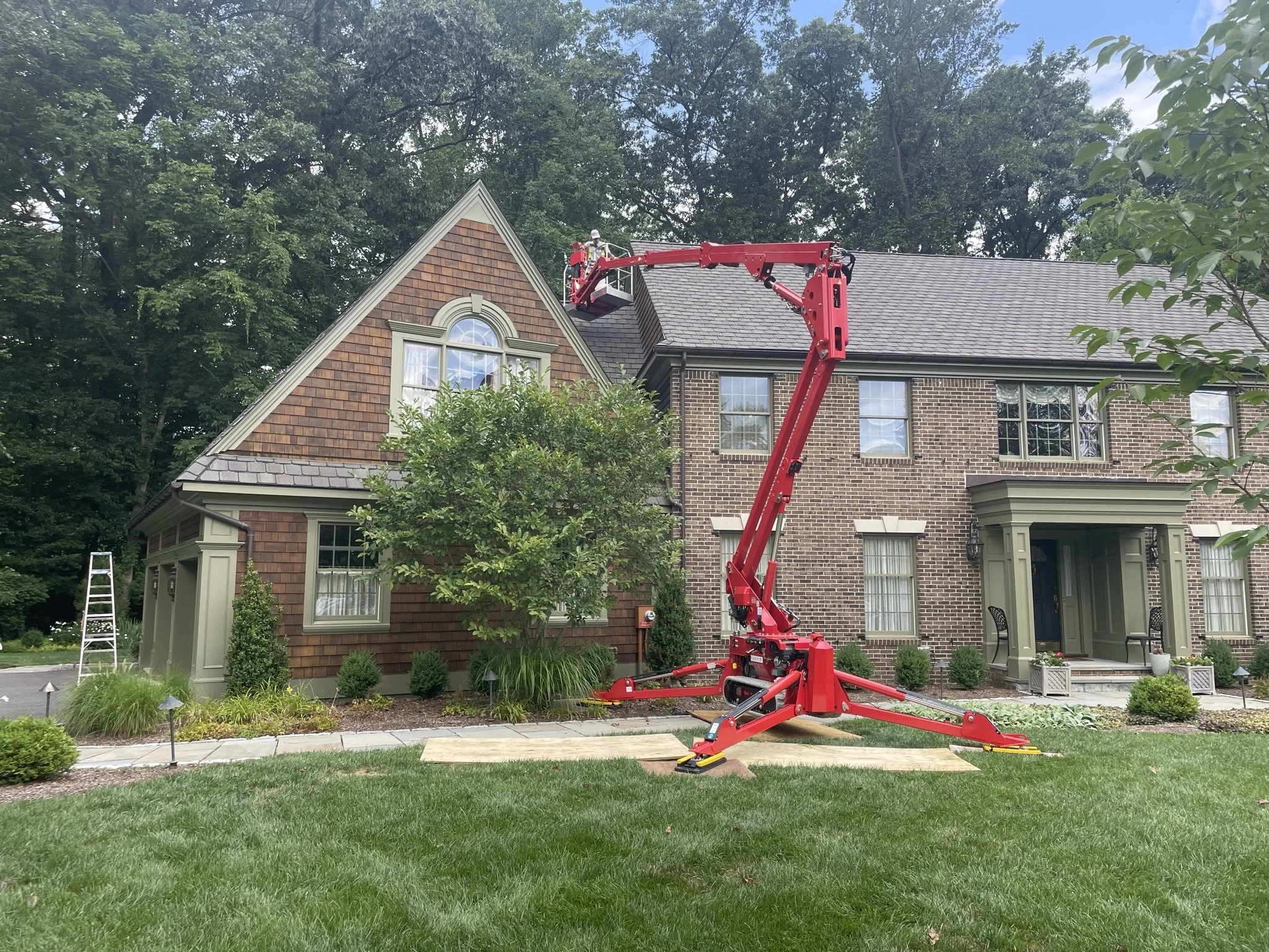 A house with a brick and shingle exterior, with a cherry picker lifted to the roof, likely for roofing or maintenance work, on a well-manicured lawn with landscaping.