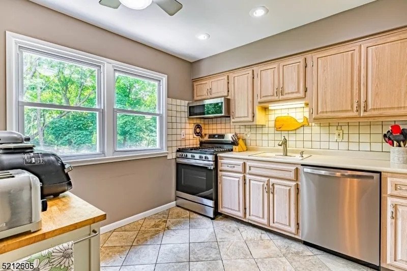 Kitchen with beige wooden cabinets, stainless steel dishwasher, stove, and microwave, white tile backsplash, window with greenery outside, tile floor, and countertop appliances.