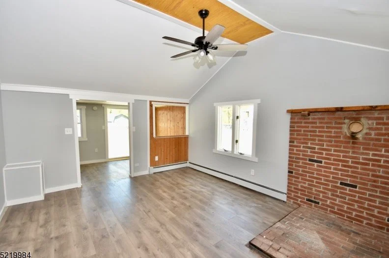 Living room with light gray walls, wood flooring, and a brick fireplace. A ceiling fan with light fixtures hangs from the ceiling, which has a wood accent section. There are two windows and a doorway leading to another room.
