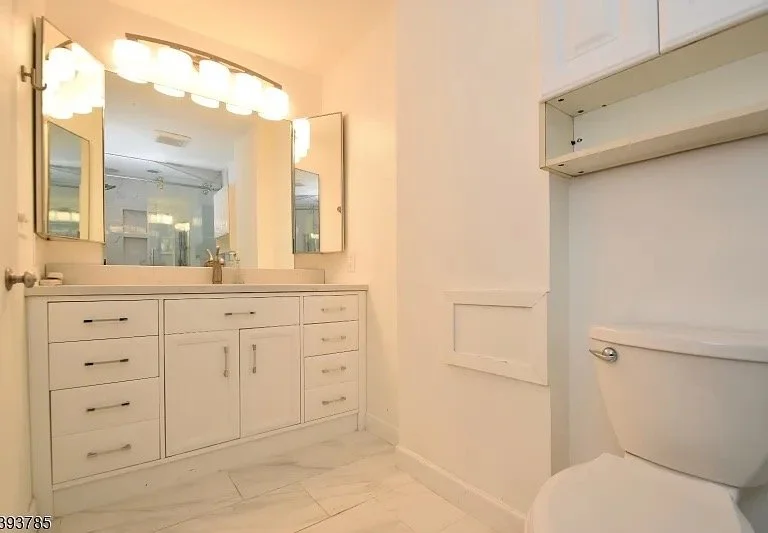 A bathroom with a white vanity, large mirror, and mounted lights above the mirror, with a toilet and open shelving on the right side.