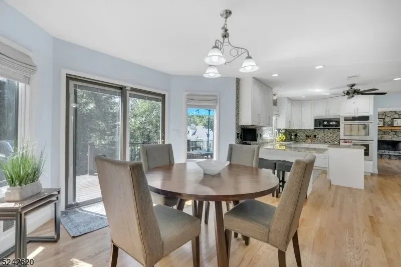 Interior view of a dining area and kitchen in a home with light wood floors, off-white cabinetry, and large windows letting in natural light.