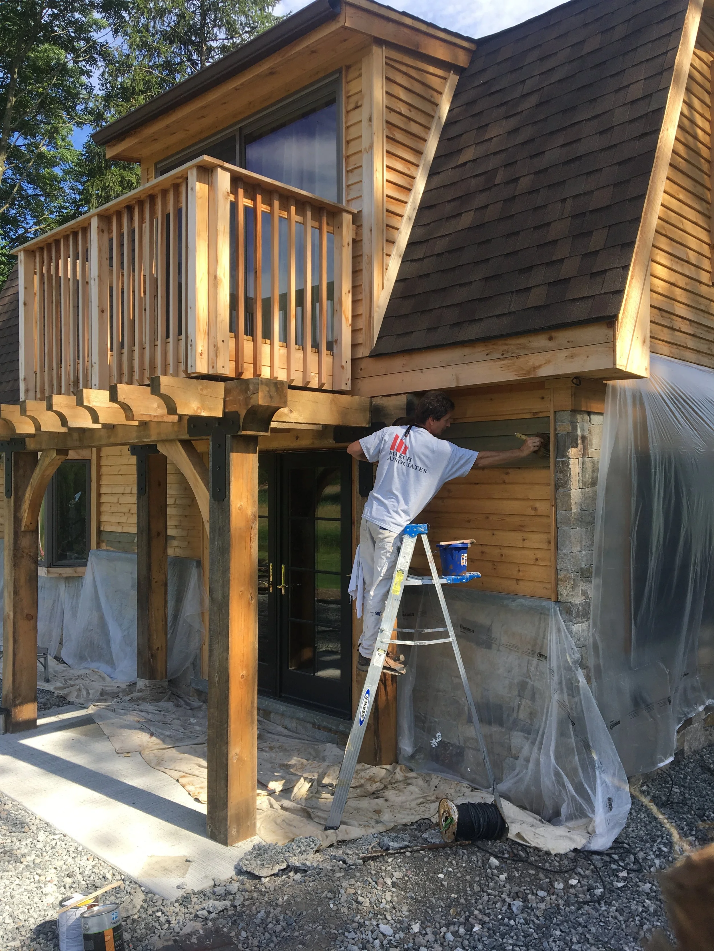 Construction worker painting the exterior wall of a two-story house with wood siding. The house has a balcony and a pitched roof. Plastic sheeting covers some areas, and construction tools and materials are on the ground.