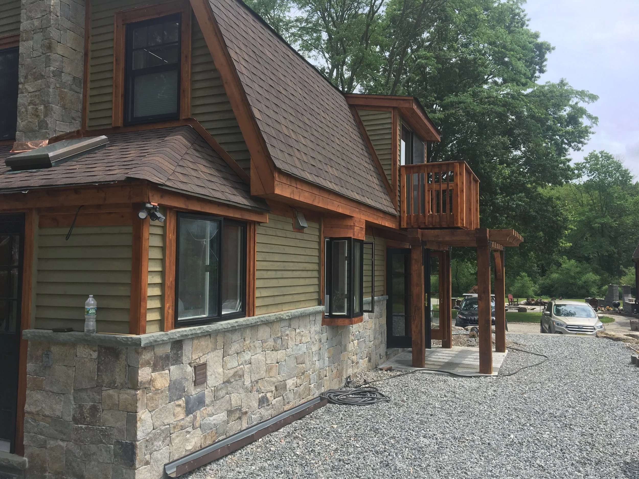 A house under construction with a stone and wood exterior, a small deck on the second floor, and cars parked nearby on a gravel driveway surrounded by green trees.