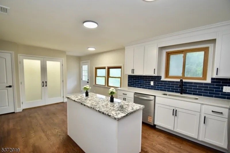Modern kitchen with white cabinets, granite island, blue subway tile backsplash, wooden window frames, and hardwood floors.