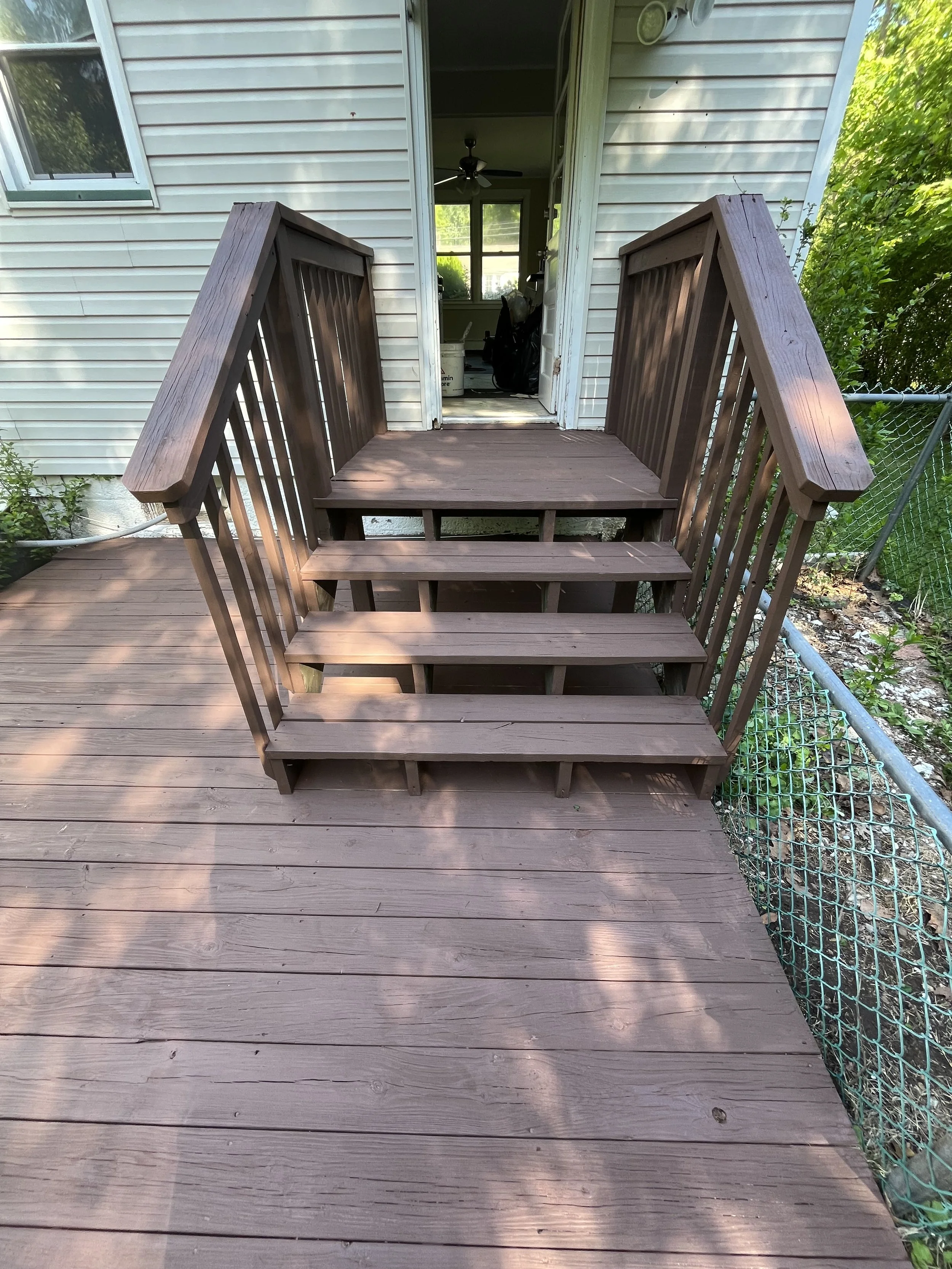 Photo of a wooden staircase leading up to a back door of a house, with a porch and surrounding greenery.
