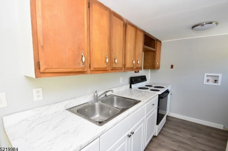 Kitchen with wooden cabinets and white marble countertop, stainless steel sink, white stove with black cooktop, and gray flooring.