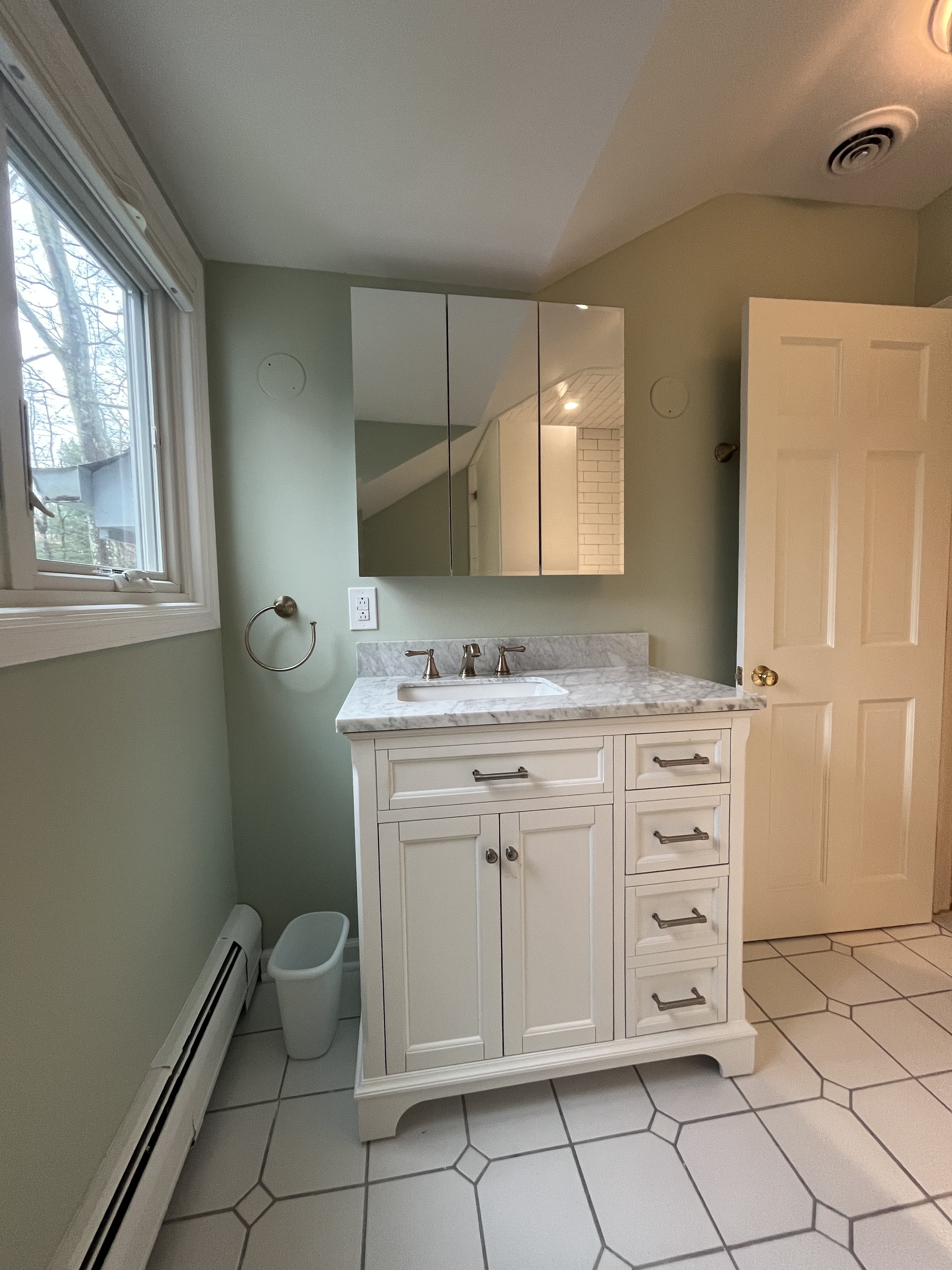 Bathroom with a white vanity with marble countertop and a sink, mirrored medicine cabinet, window, tiled floor, door, and a small trash can.