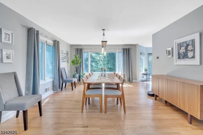 Bright dining room with a wooden table, six chairs, large windows with curtains, hardwood flooring, framed art on walls, and a sideboard.