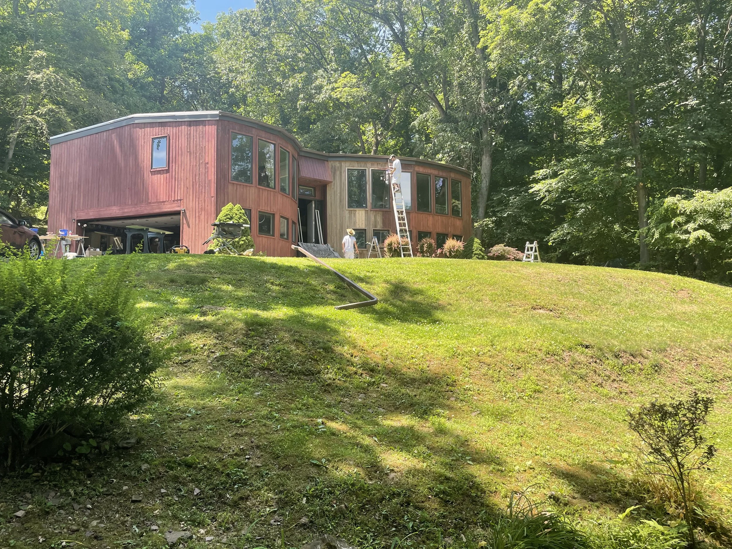 People working on the exterior of a two-tone, red and natural wood, modern, curved house situated on a grassy hill surrounded by trees.
