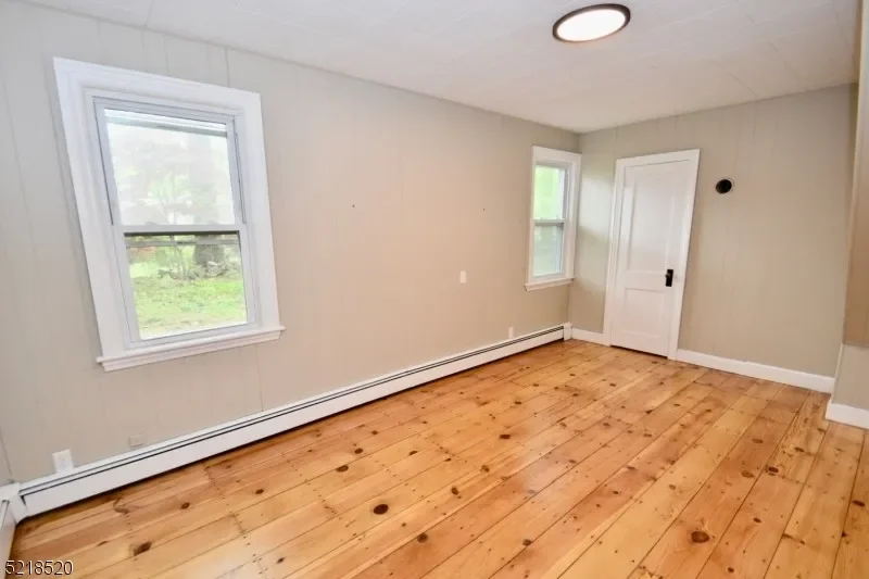 Empty room with beige walls, wooden floor, two windows, a white door, and a ceiling light fixture.