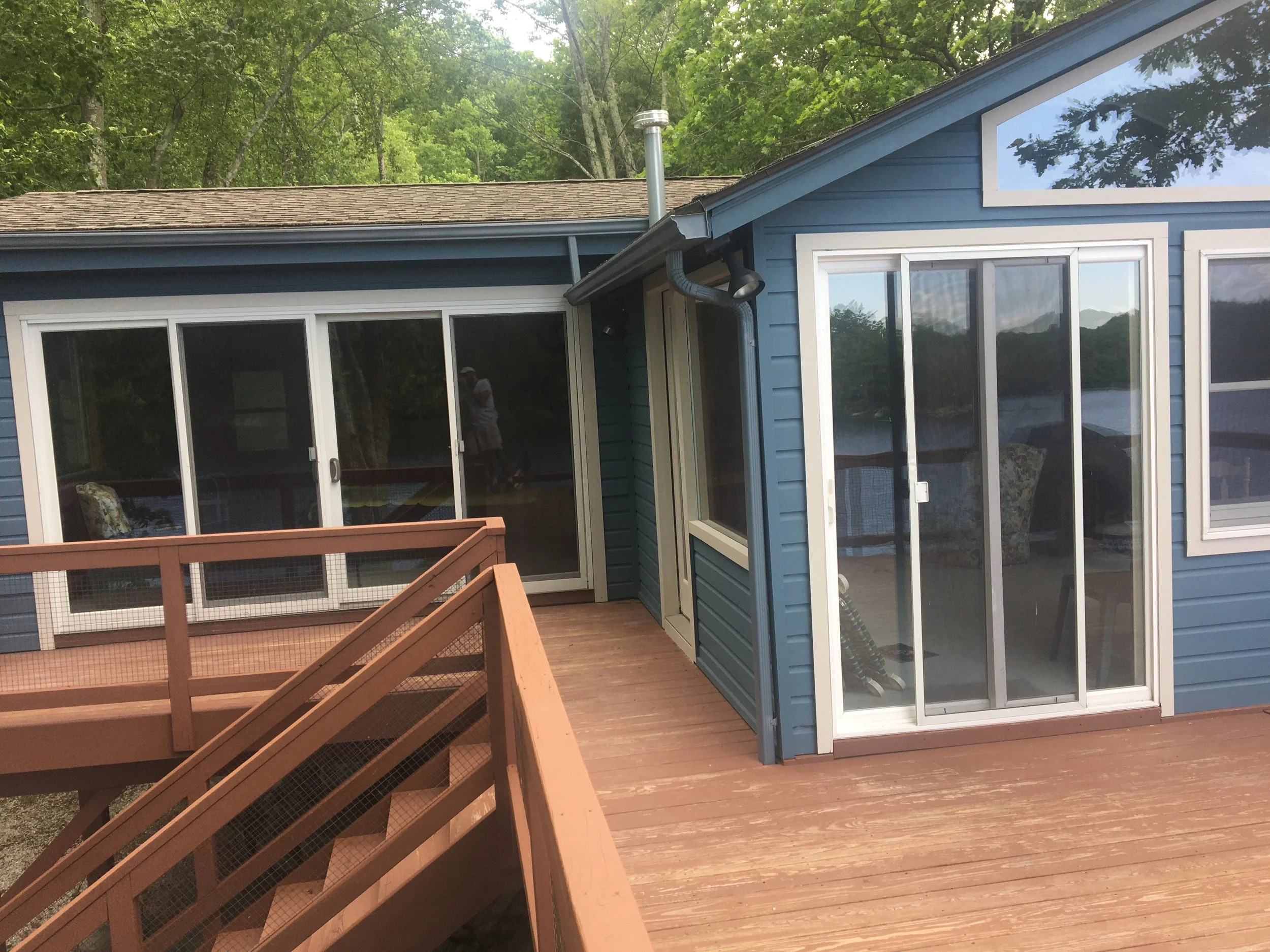 Photo of a blue house with large sliding glass doors leading to a wooden deck outside, with trees and a body of water visible in the background.