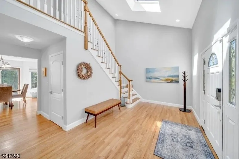Entryway of a house with hardwood floor, white walls, and a staircase with a wooden handrail. A coat rack, a bench, wall decor, and a painting are visible. Sunlight enters through the front door with glass windows.