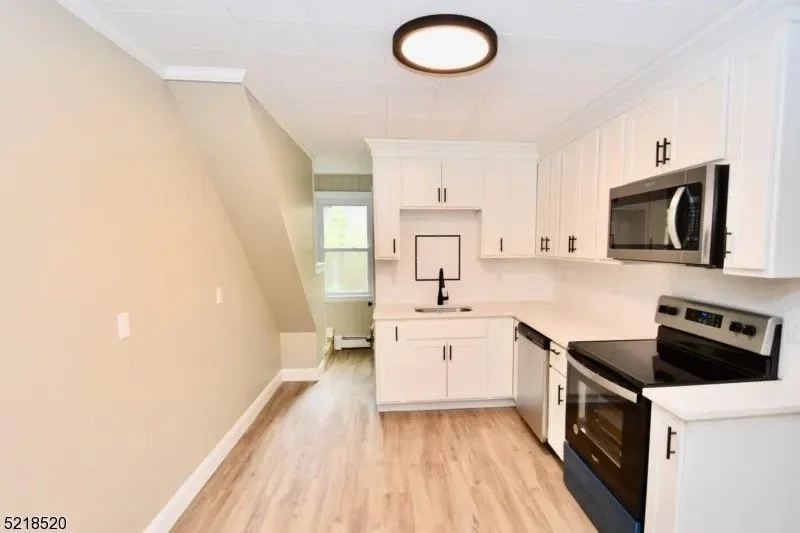 Photo of a modern kitchen with white cabinets, black appliances, a small sink, and a window. The room has light-colored walls, wood floors, and a ceiling light.