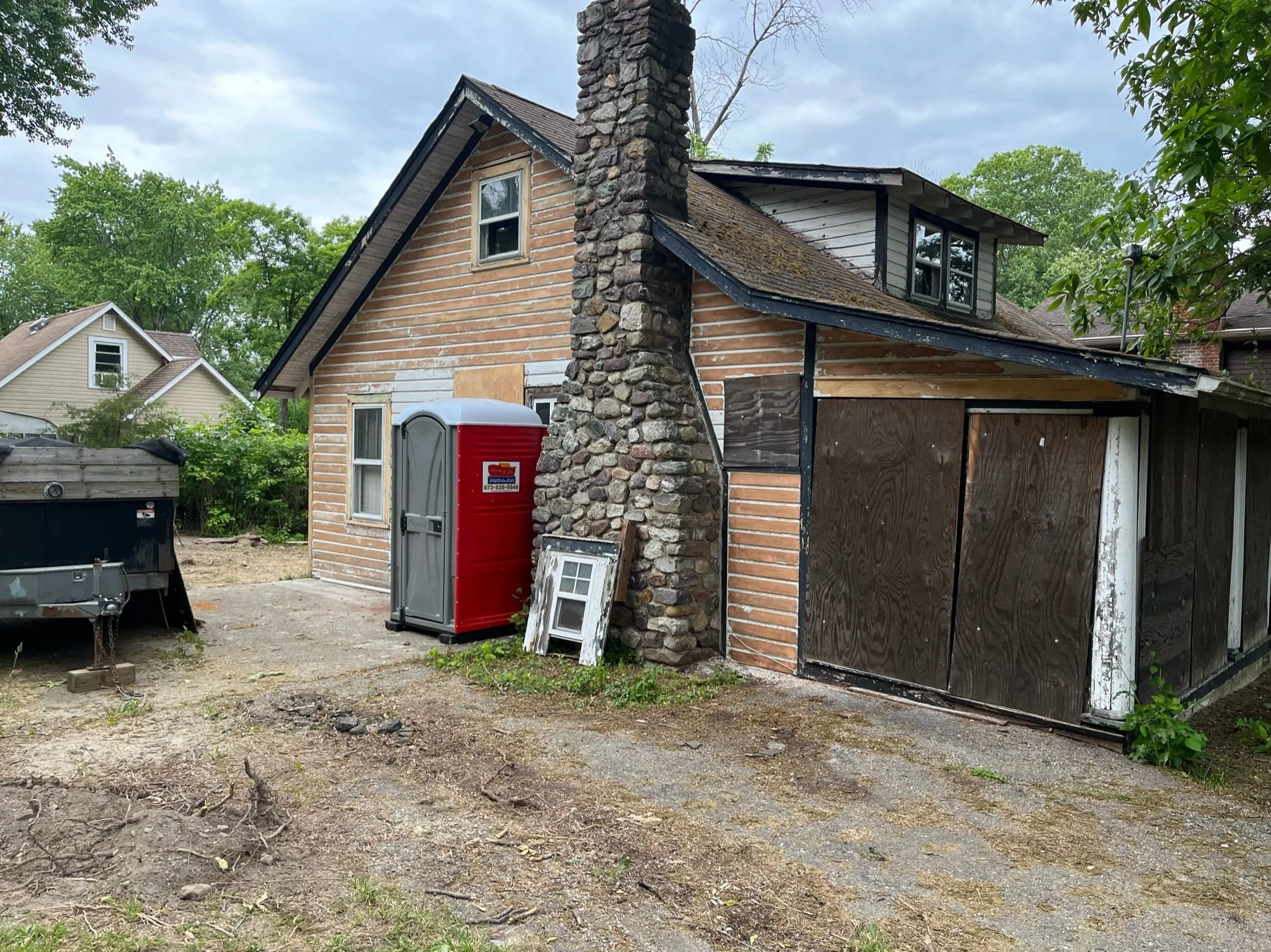 A partially renovated two-story house with a brick chimney, wooden siding, and boarded-up garage door. A red portable toilet and a small window frame are outside on a gravel driveway. Green trees and neighboring houses are visible in the background.