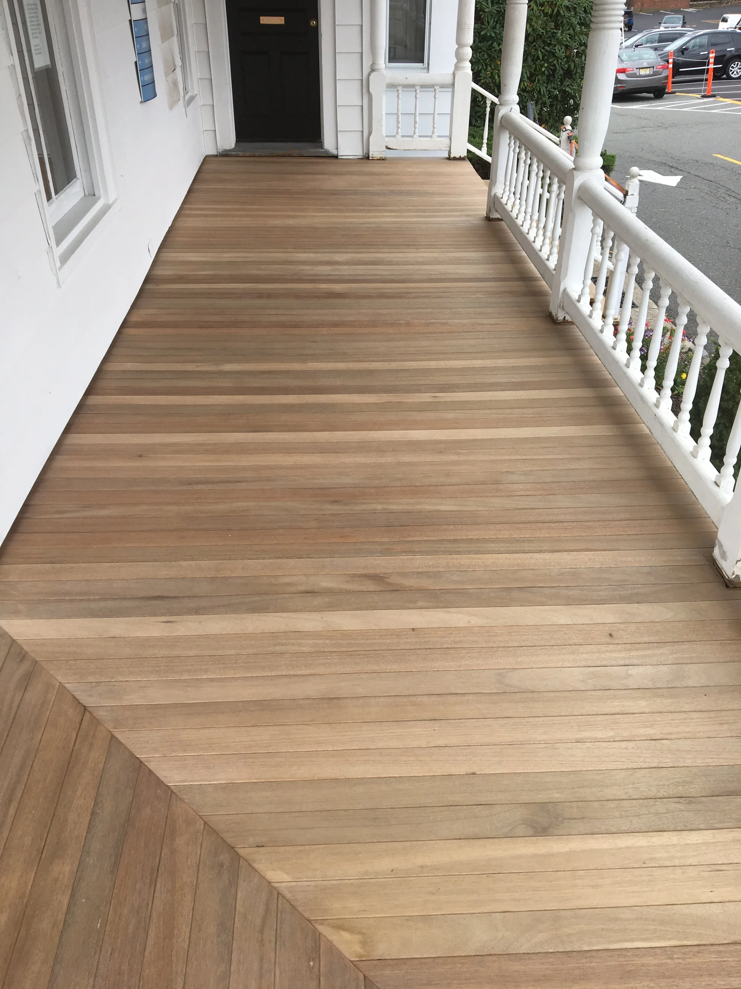 A front porch with a wooden deck and white railing, leading to a black door, with parked cars visible on the street in the background.